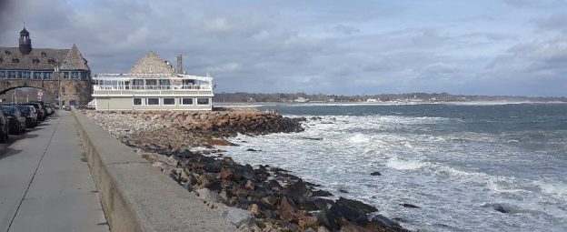 A picture of stone towers on each side of a road with a bridge connecting them. The sea is off to the right.